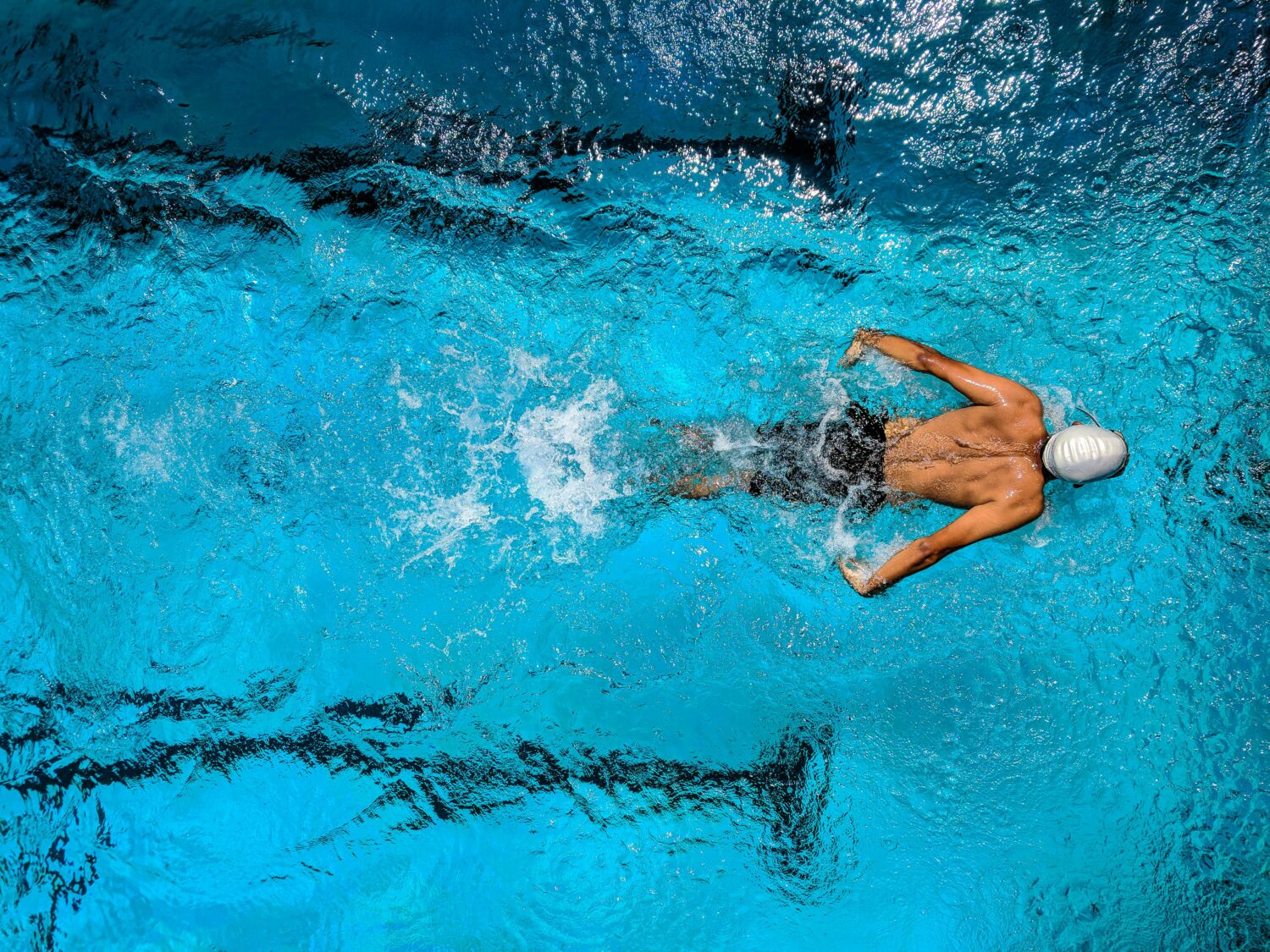 over head camera shot of a man in a swimming pool wearing black swimming shorts and a grey swimming cap doing the butterfly stroke