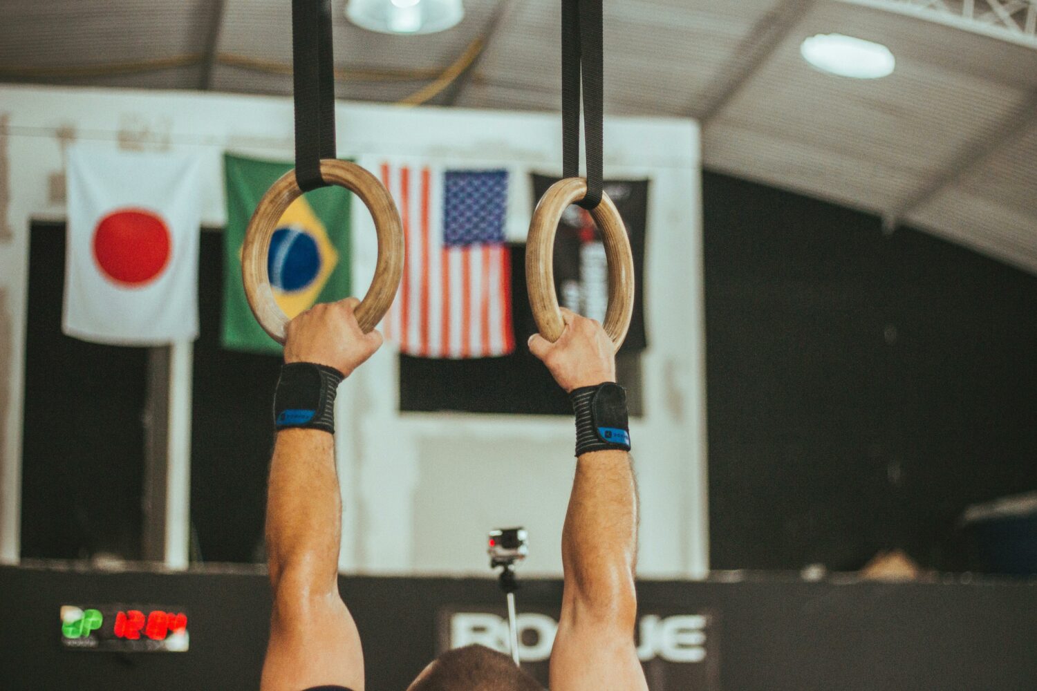 A man holding on to the gymnastics rings with the Japan, Brazil and USA flag in the background